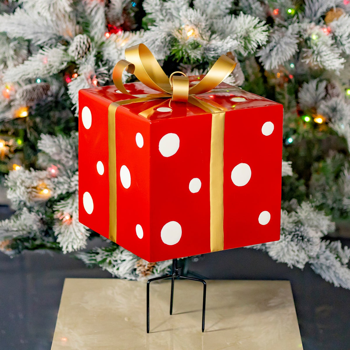 Angled view of the red polka-dot gift box, gold ribbon bow and black stake against a Christmas tree.