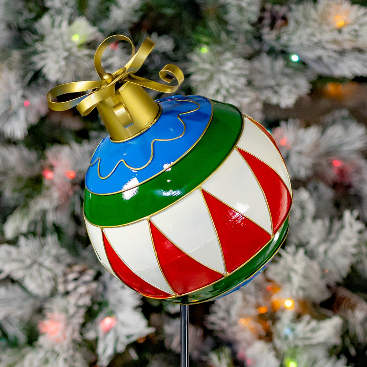 A glossy striped ornament with a gold bow and blue and green bands against a snowy tree.