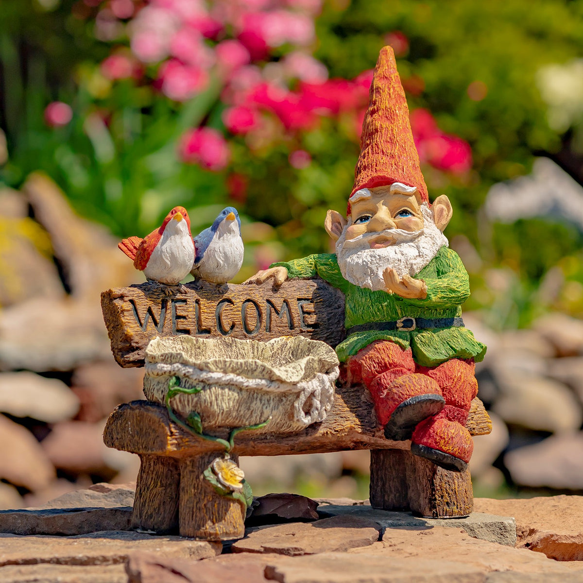 Garden ornament showing the gnome sitting on a wooden bench beside a birdbath and two colorful birds.