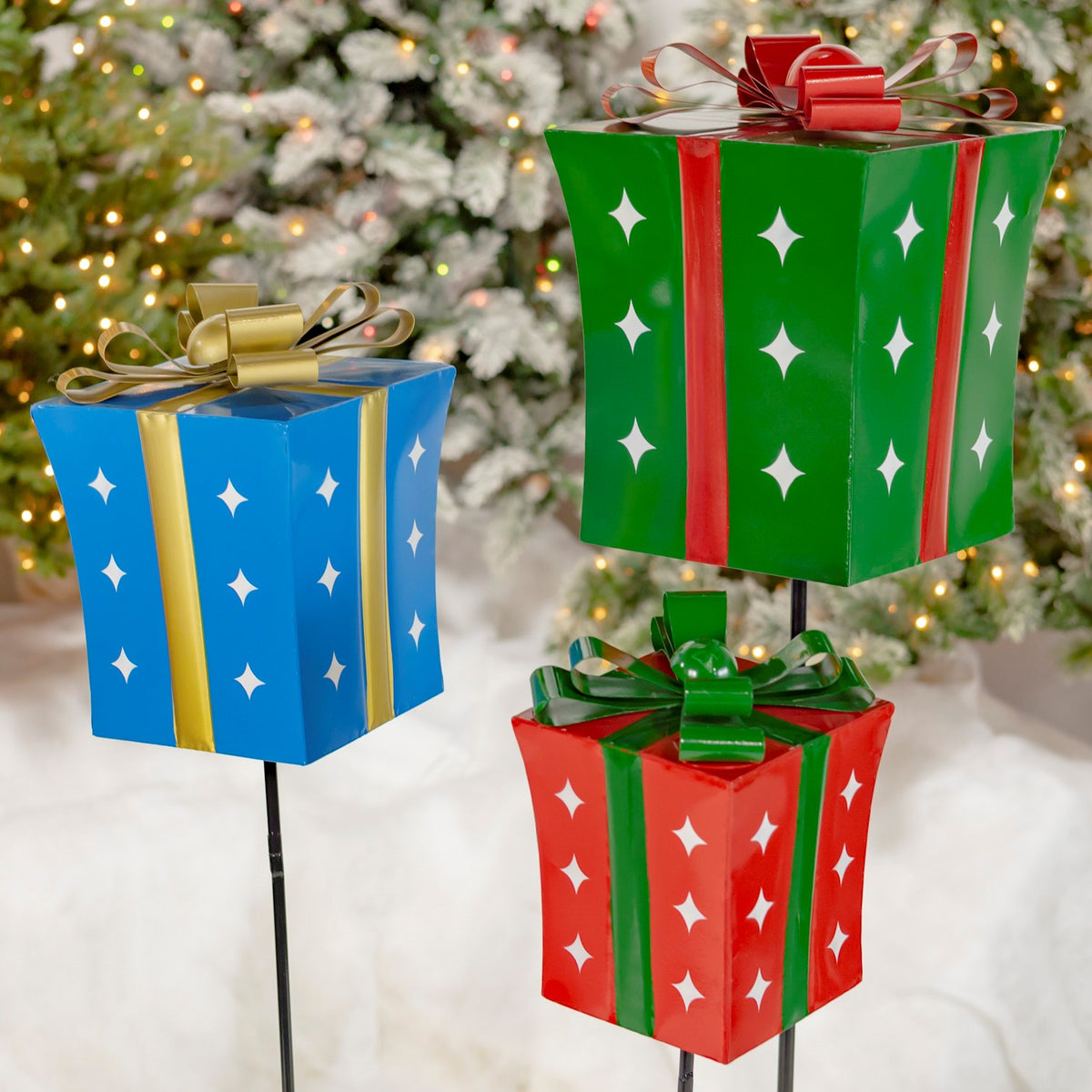 Blue, green, and red gift-box stakes with bows and white star cutouts against snowy branches.