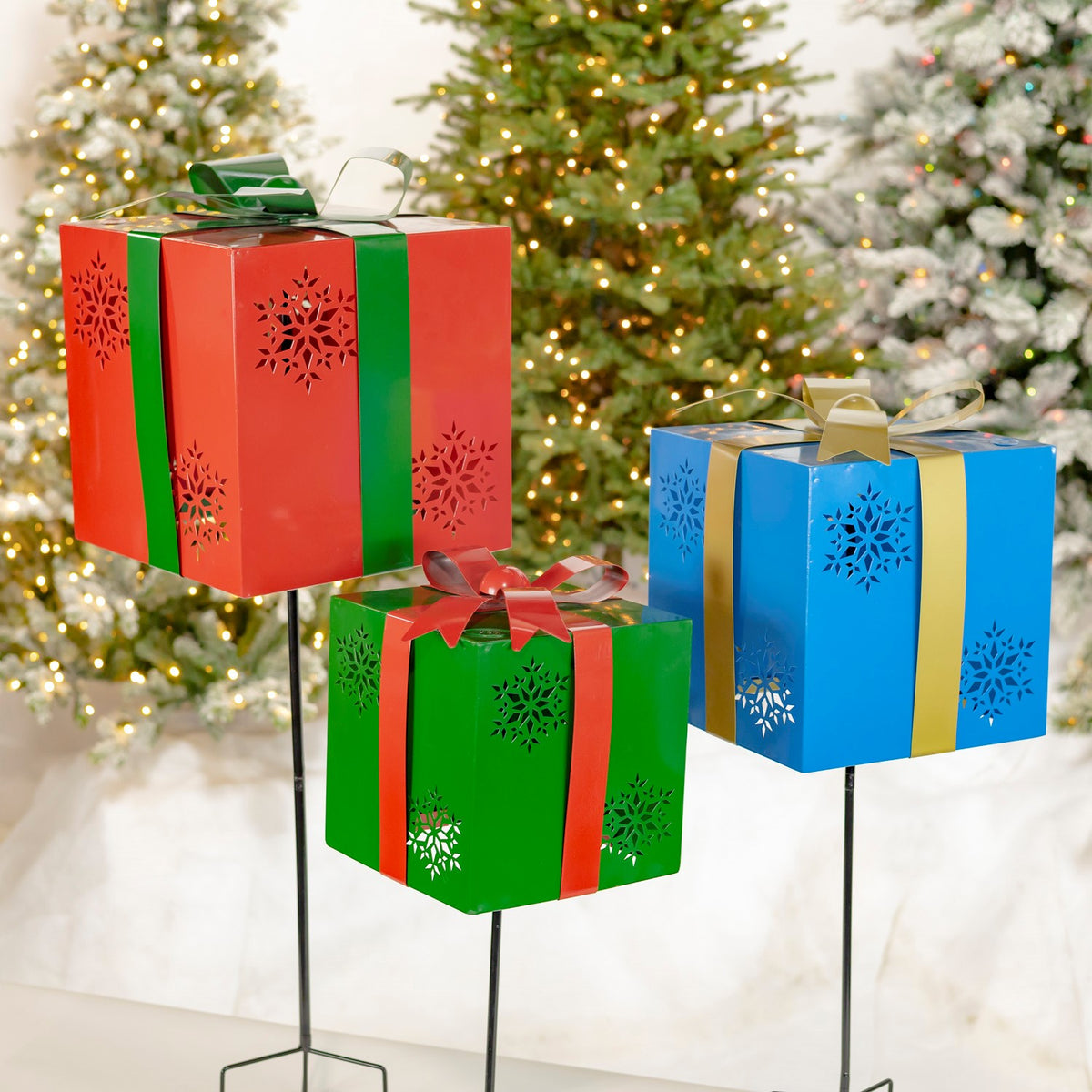 Three colorful gift-box stakes with snowflake cutouts viewed against a lit Christmas tree