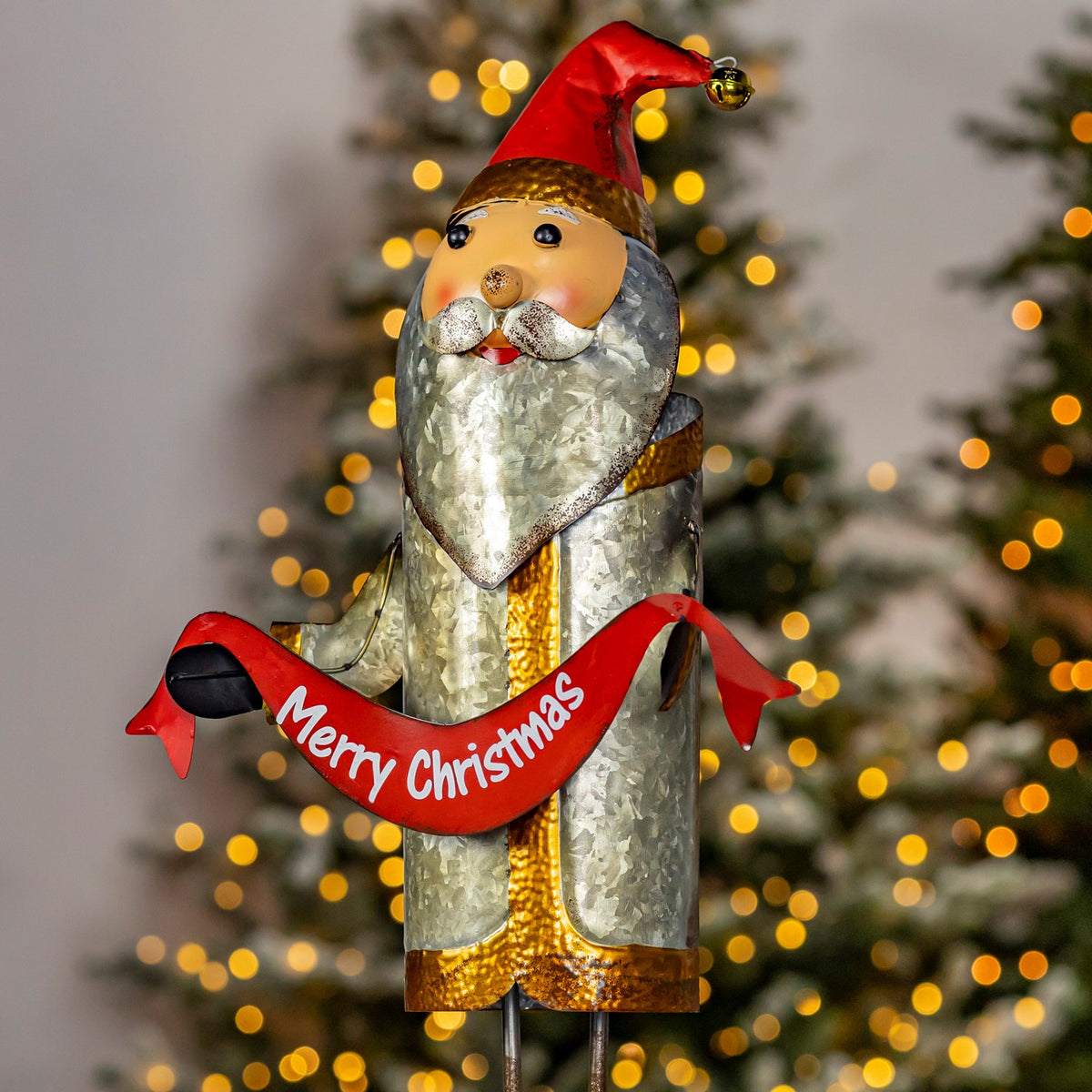 Front view of iron Santa holding a red Merry Christmas sash by a tree