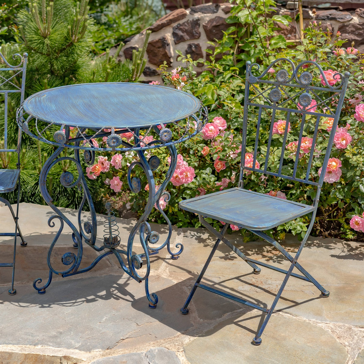 Blue metal bistro table and folding chairs arranged on a garden patio among flowers.