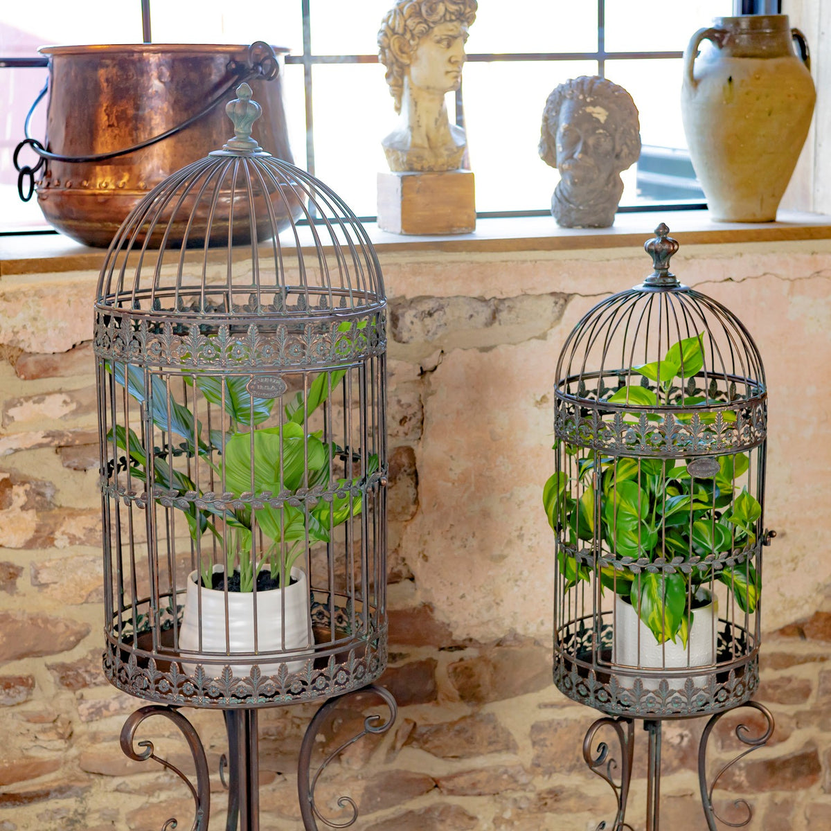 Two dark iron birdcage planters holding leafy green plants beside a stone wall.