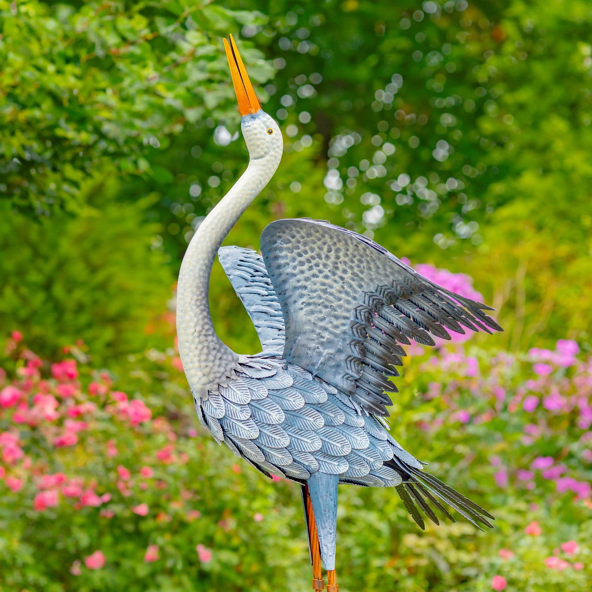 Blue-grey heron figurine with spread wings shown full-length in a flower garden