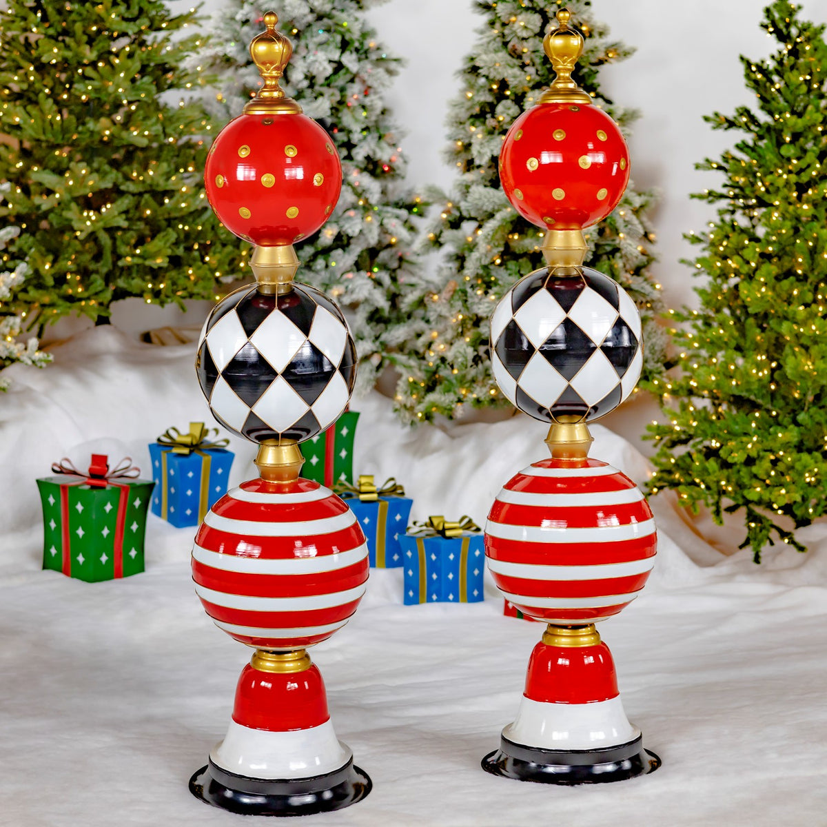 Two matching ornament towers stand side by side among wrapped gifts and snowy Christmas trees.