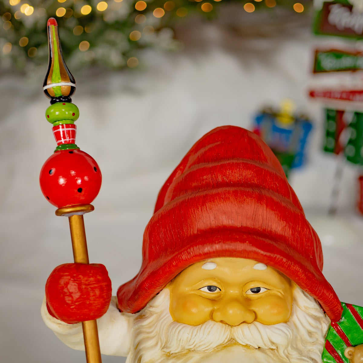 Close-up of a gnome’s face, red hat, white beard and hand gripping a staff topped with colorful ornaments.