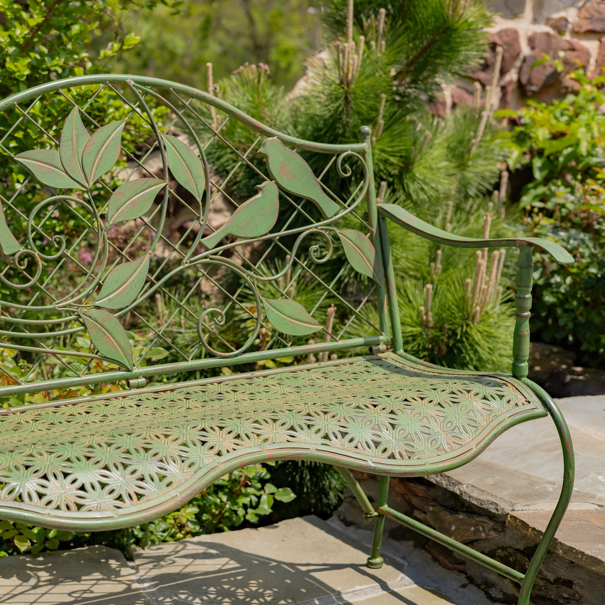 A green metal garden bench with leafy scrollwork and an ornate perforated seat stands beside a stone path.
