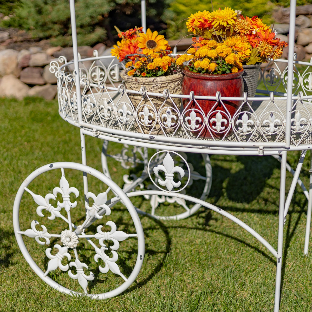 Close view of the white cart’s fleur-de-lis rail, flower pots, and ornate spoked wheel.