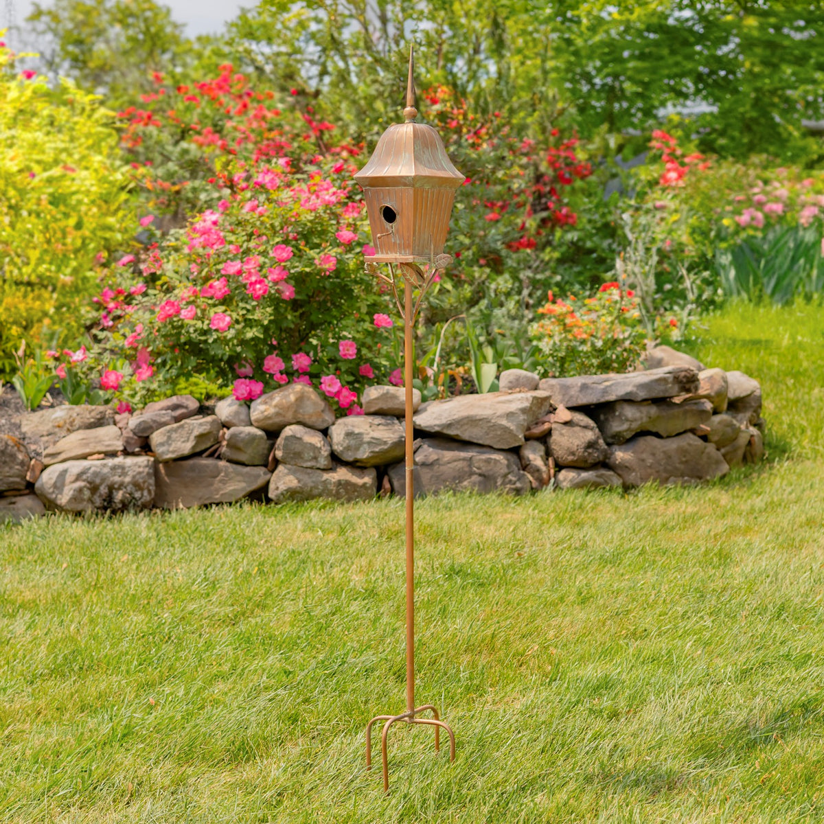 Full birdhouse stake standing in grass, with a three-pronged base and a stone garden border behind it.