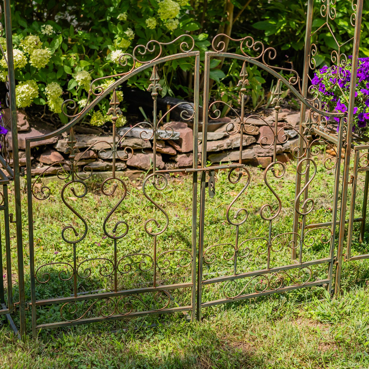 Garden archway's ornate metal side panels stand on grass in front of a low stone border and flowering plants.