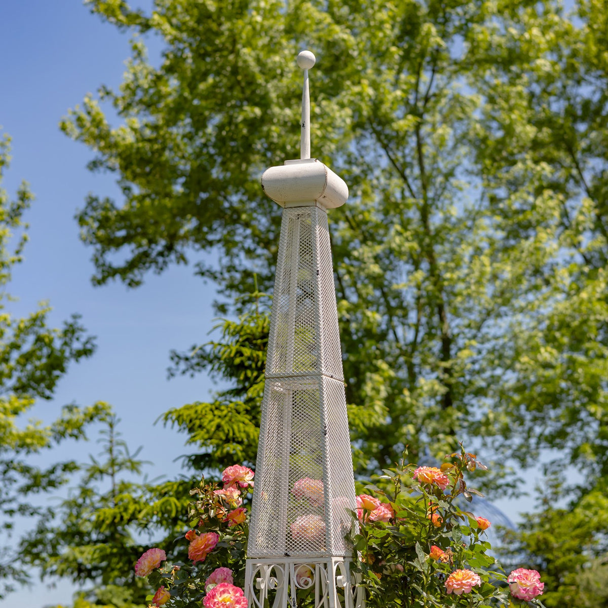Close view of the white stand’s tapered mesh tower and finial against trees and pink flowers.