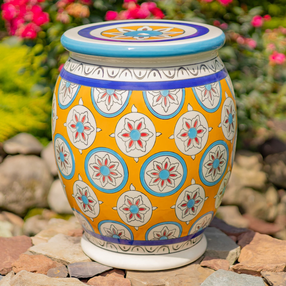 Yellow stool with repeating floral medallions, shown on a stone patio among flowers.
