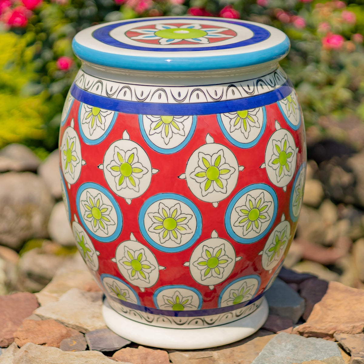A colorful ceramic stool with a rounded red floral body, blue accents, and a decorated top stands on rocks.