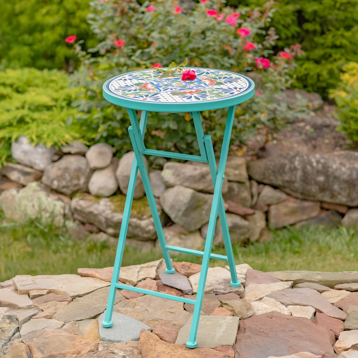 Folding mosaic plant stand with a small pink flower resting on its patterned top, set on a stone path.