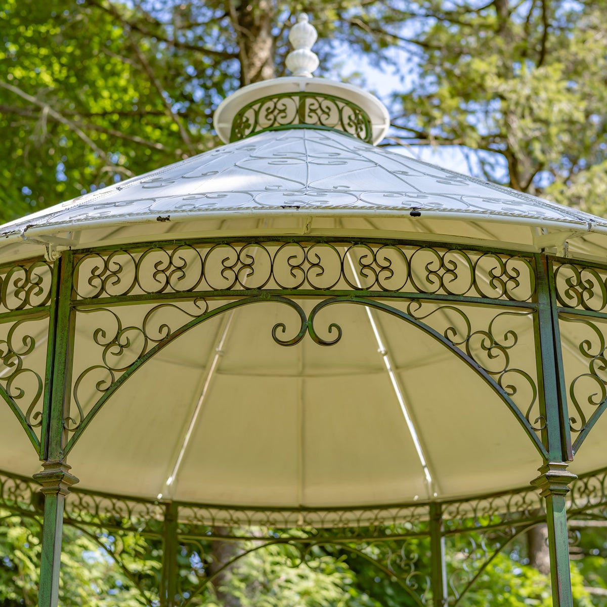 Upward view of the gazebo’s pale roof, finial, and scrolling metalwork beneath the eaves.