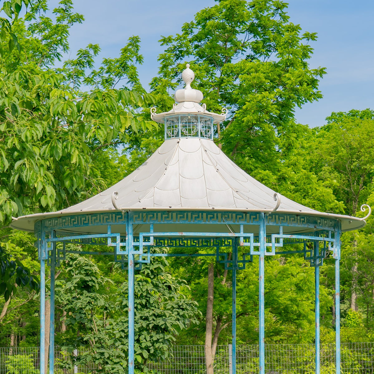 Round gazebo viewed from the front, showing its pale conical roof, decorative finial and open iron framework.
