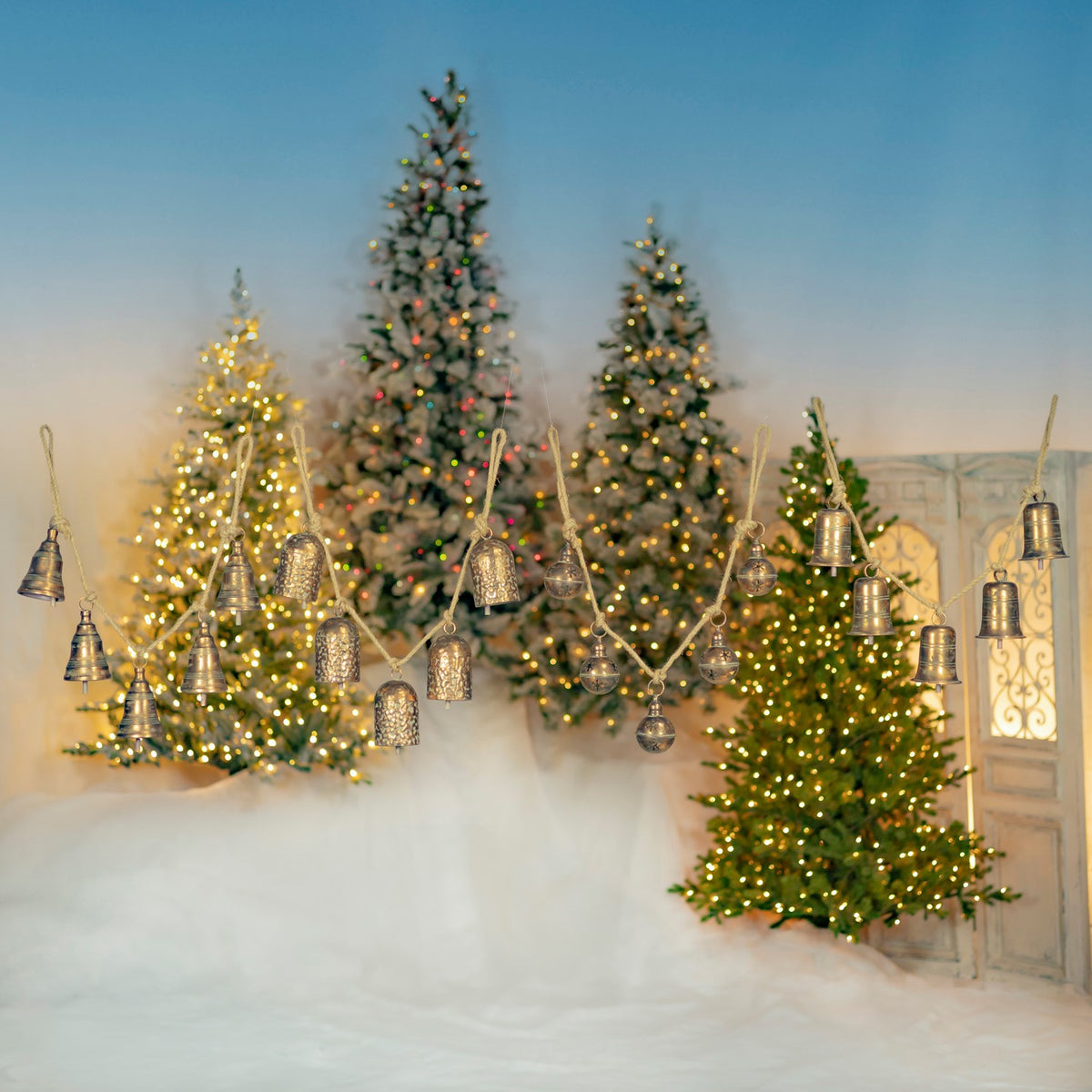 Several styles of hanging metal bells are displayed on ropes among illuminated Christmas trees.