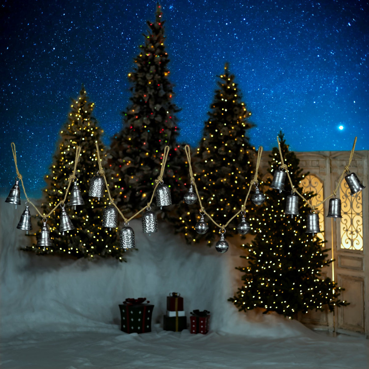 A strand of assorted silver bells hangs across a snowy scene with lit Christmas trees.