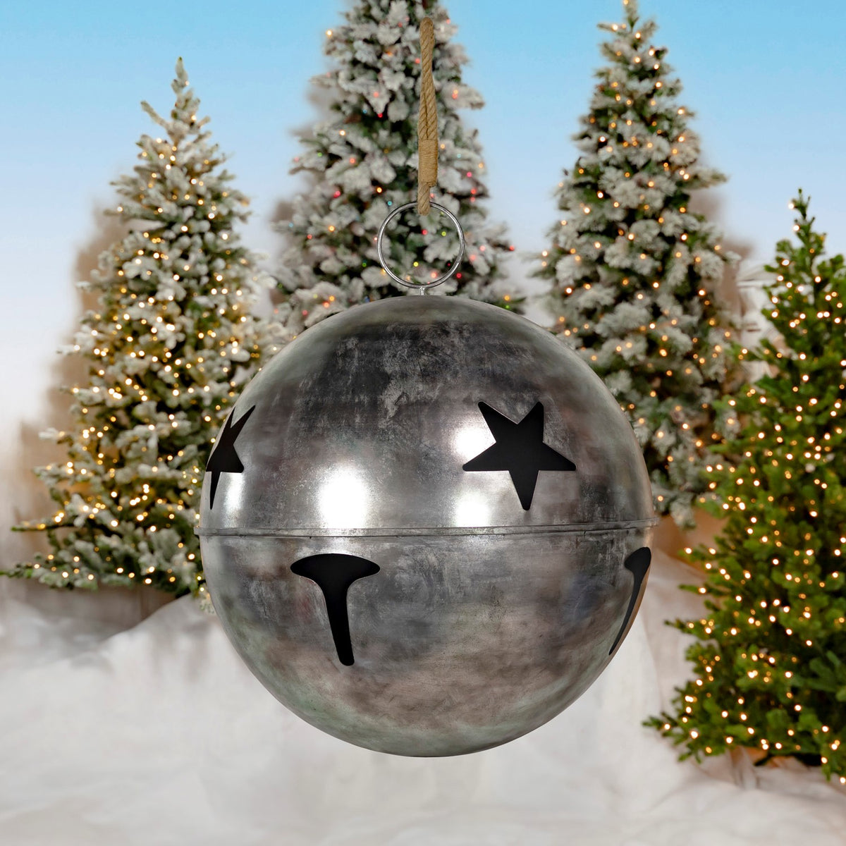 Large silver bell hanging by a cord, with star cutouts and dark openings against lit Christmas trees.