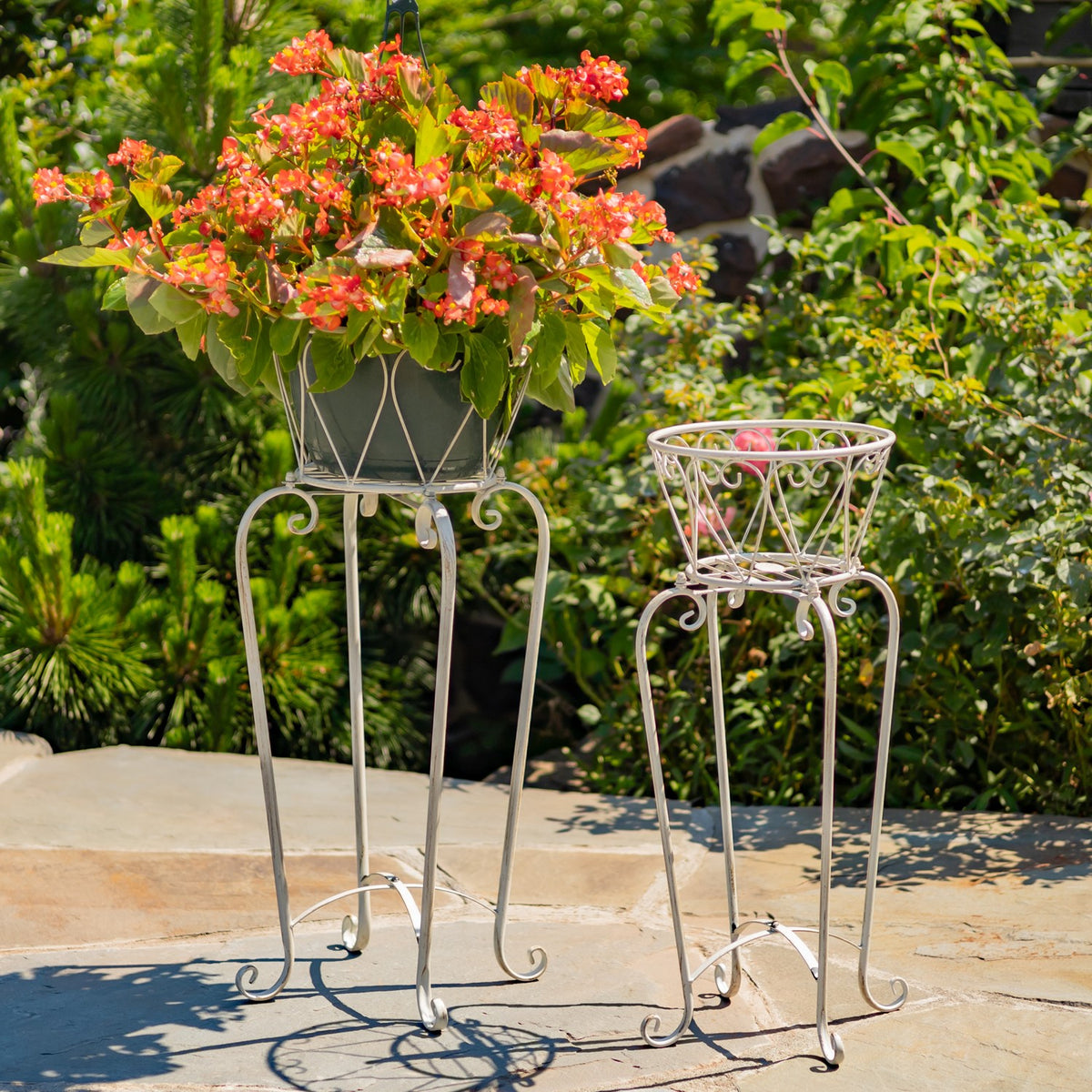 Pair of white metal basket stands on a stone patio, one holding a flowering plant.