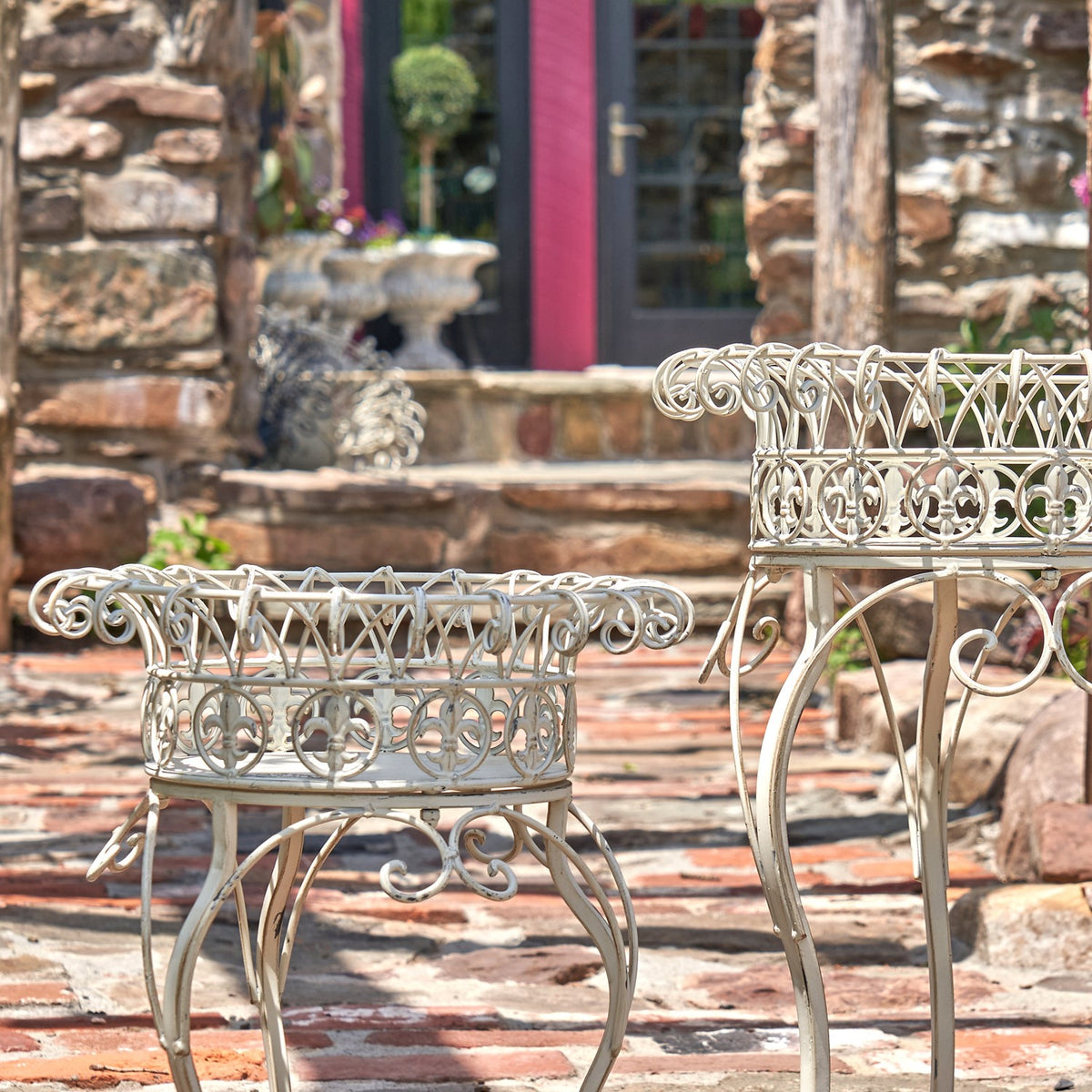 A smaller light-colored plant stand in front of a taller matching stand, showing their scrollwork baskets and legs.