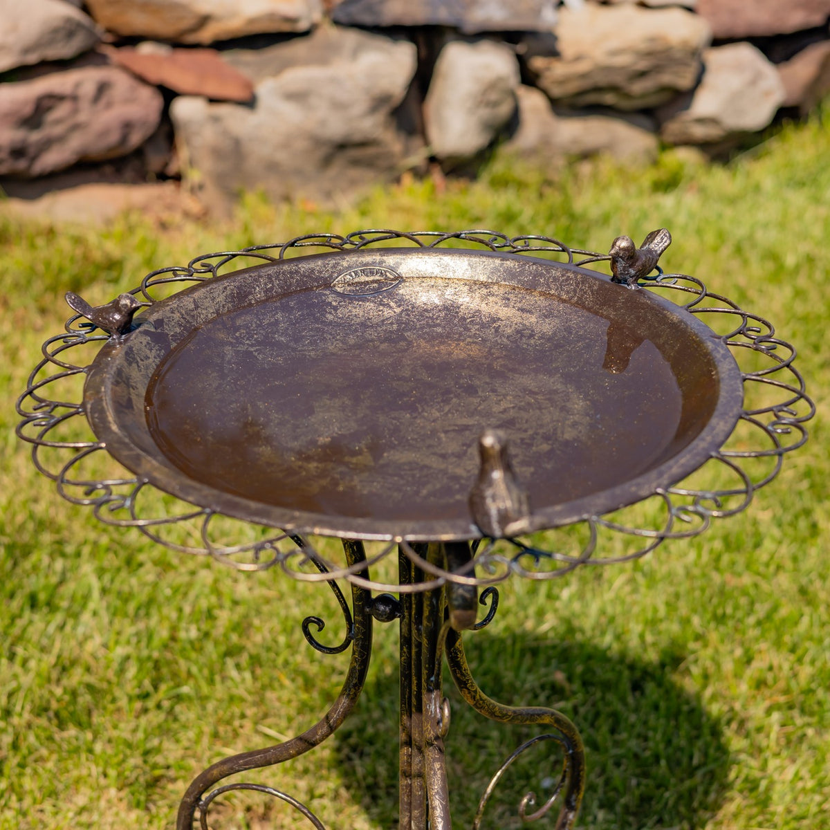 Round birdbath seen from above, showing its decorative looped rim, bird figures, and curled pedestal legs.