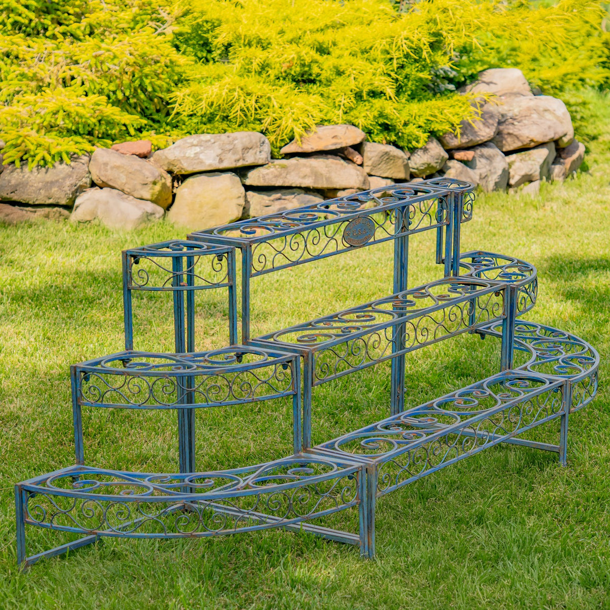 Blue tiered plant stand shown from the opposite side on grass beside a low stone wall.