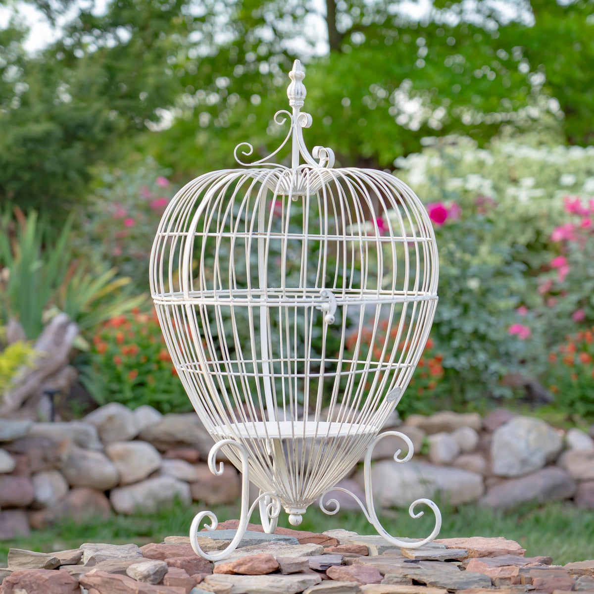 A pale wire cage plant stand with curled feet and a decorative finial stands among garden rocks.