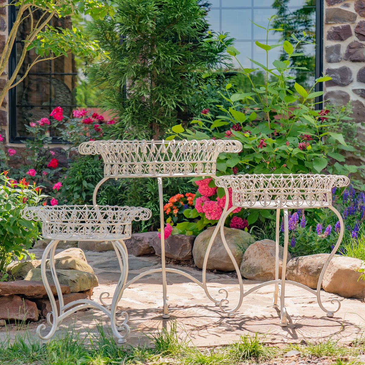 Pale wire basket stands grouped on a patio, with flowering shrubs and a stone wall behind them.