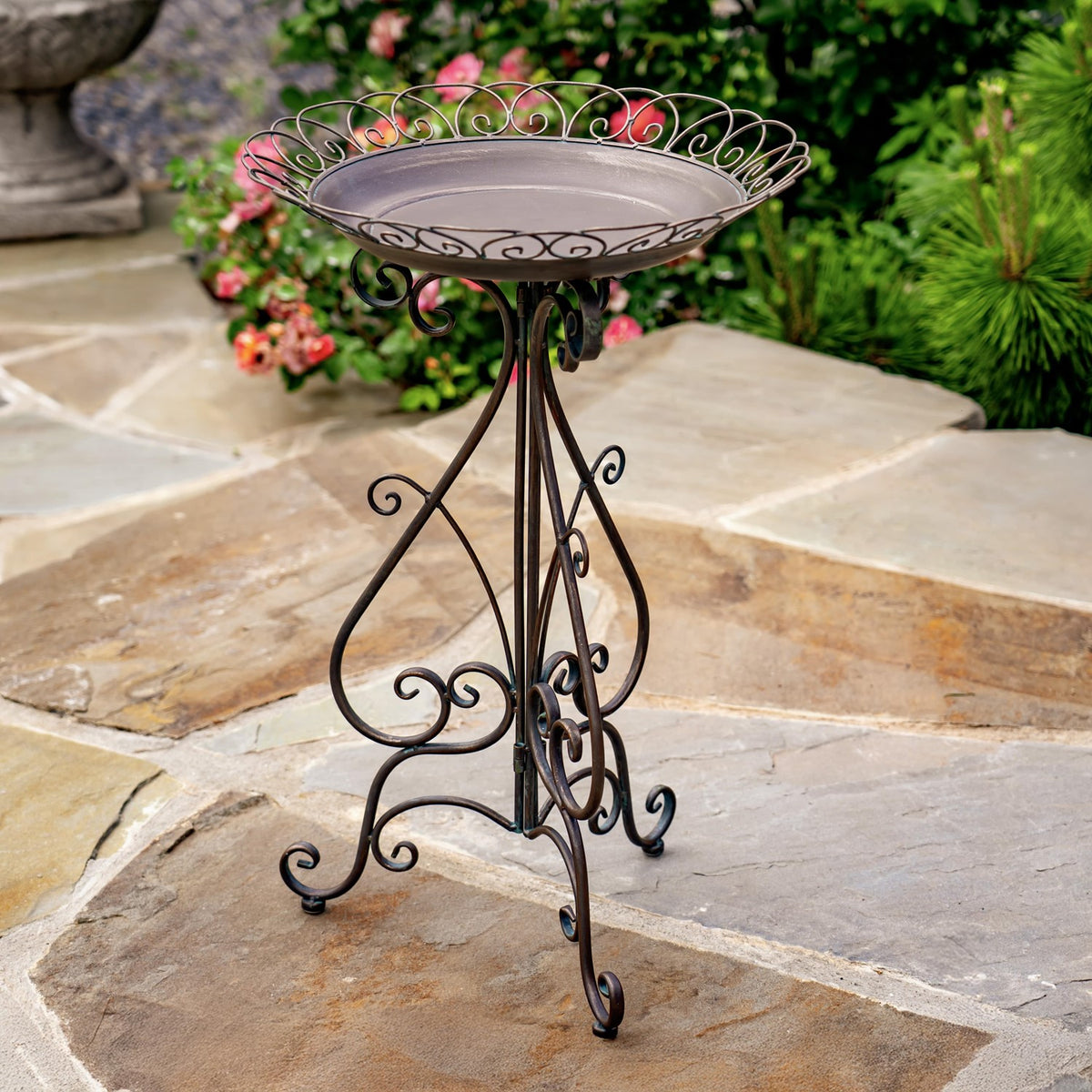 Ornate round birdbath with a shallow bowl and curled iron stand on a stone patio