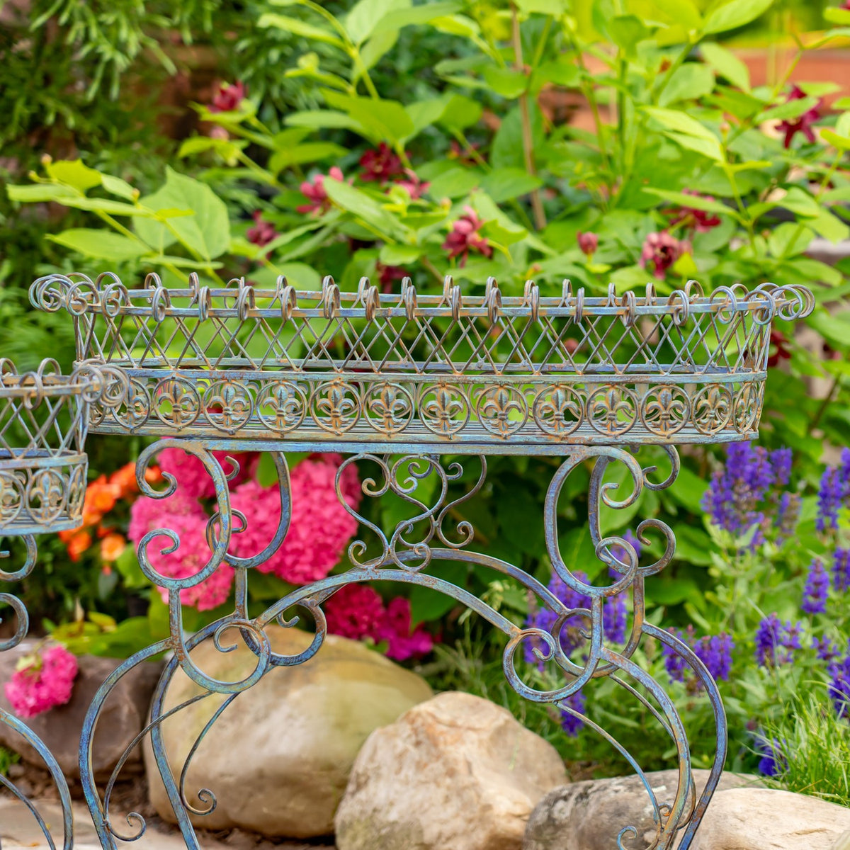 Ornate oval iron plant stand with curled scrollwork legs, set among rocks and colorful garden flowers.