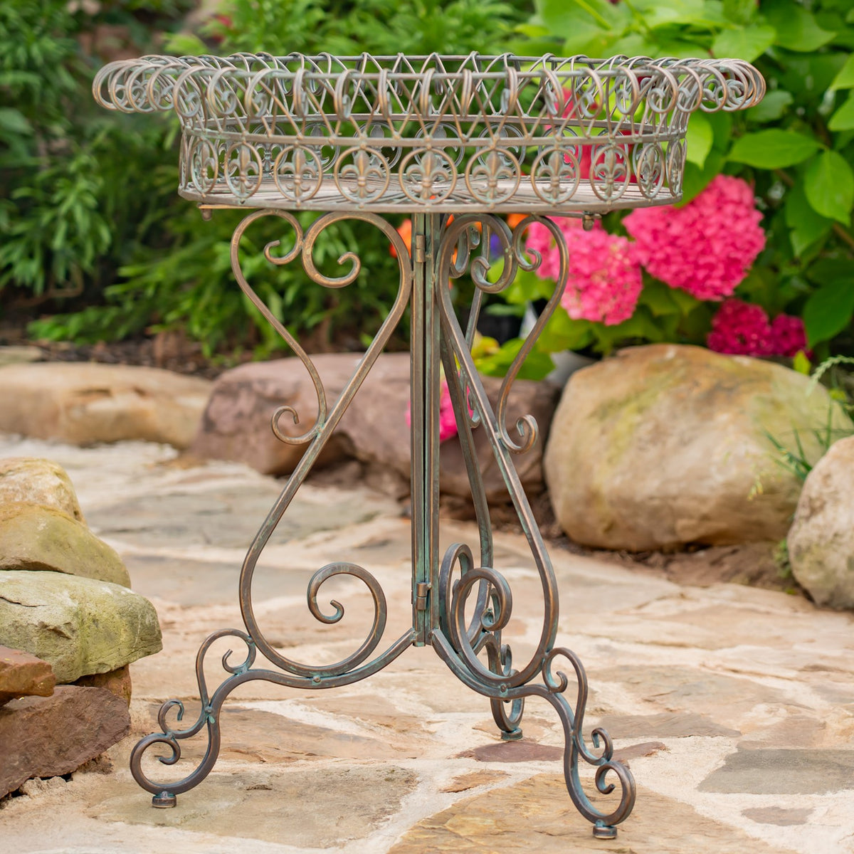 An ornate round wire plant stand with a shallow basket top and curled metal legs outdoors.