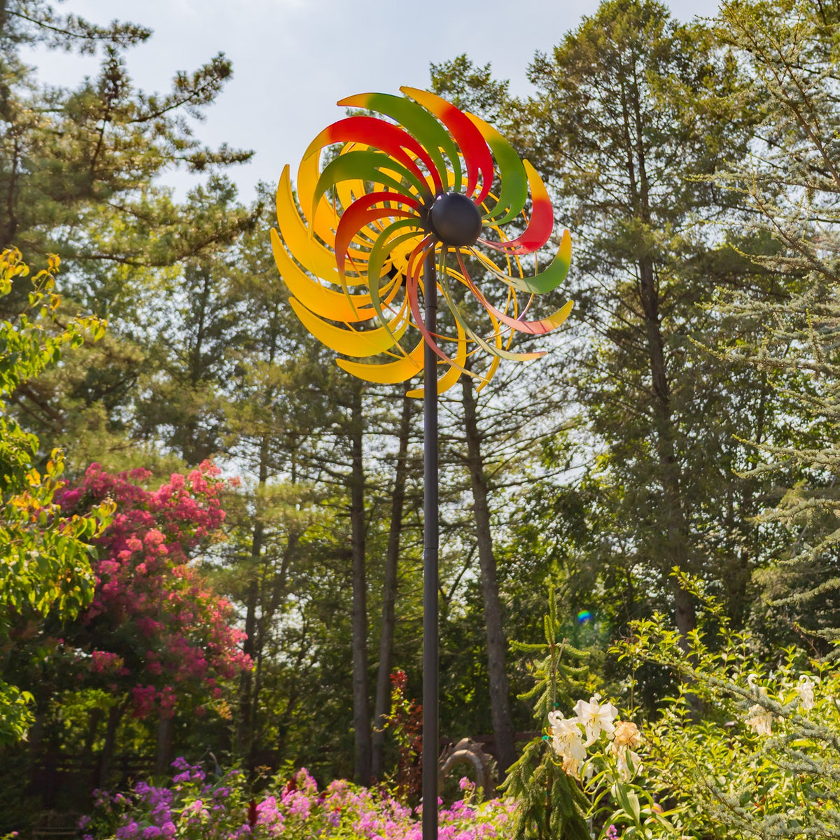 A tall multicolored windmill stake rises above flowers and greenery in a landscaped garden.