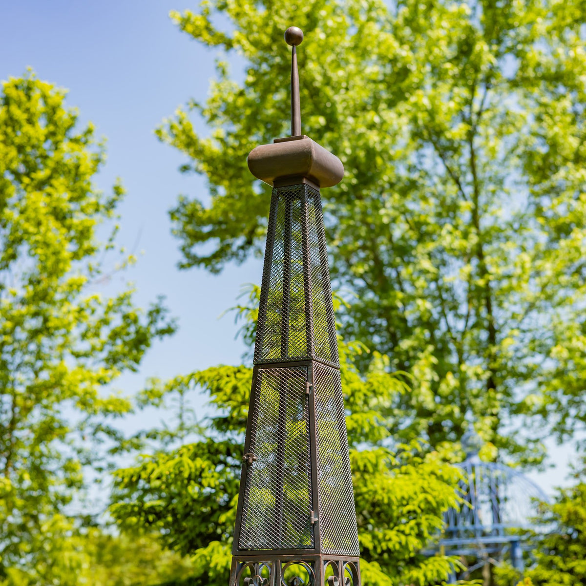 Tall, narrow iron tower plant stand with mesh panels and an ornamental finial, photographed among leafy trees.