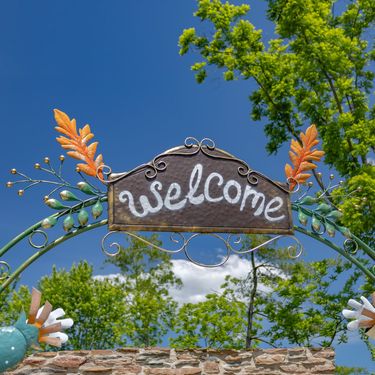 Close-up of the curved green arch and hanging welcome sign, with orange leaf accents against a blue sky.