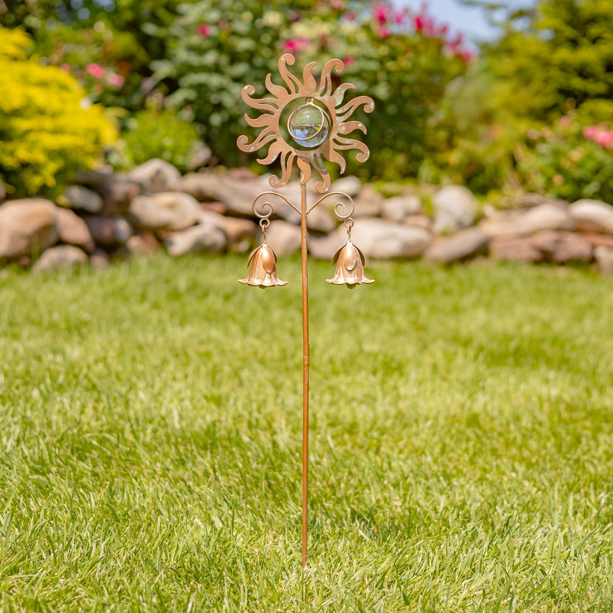 A slender sun-and-bell garden stake stands upright in a grassy garden among flowers and stones.
