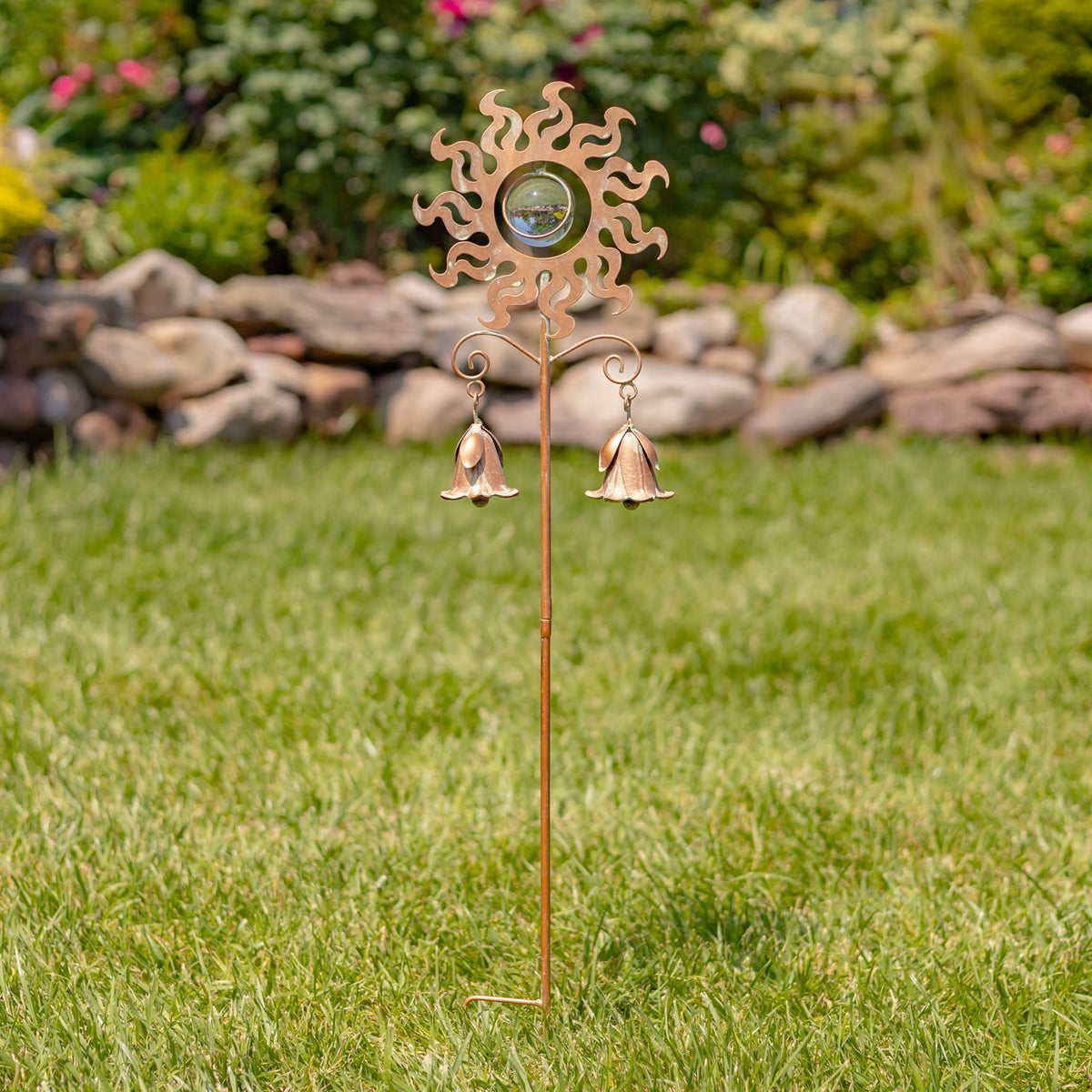 A slender garden stake stands in grass, topped with a sun ornament and two dangling bells.