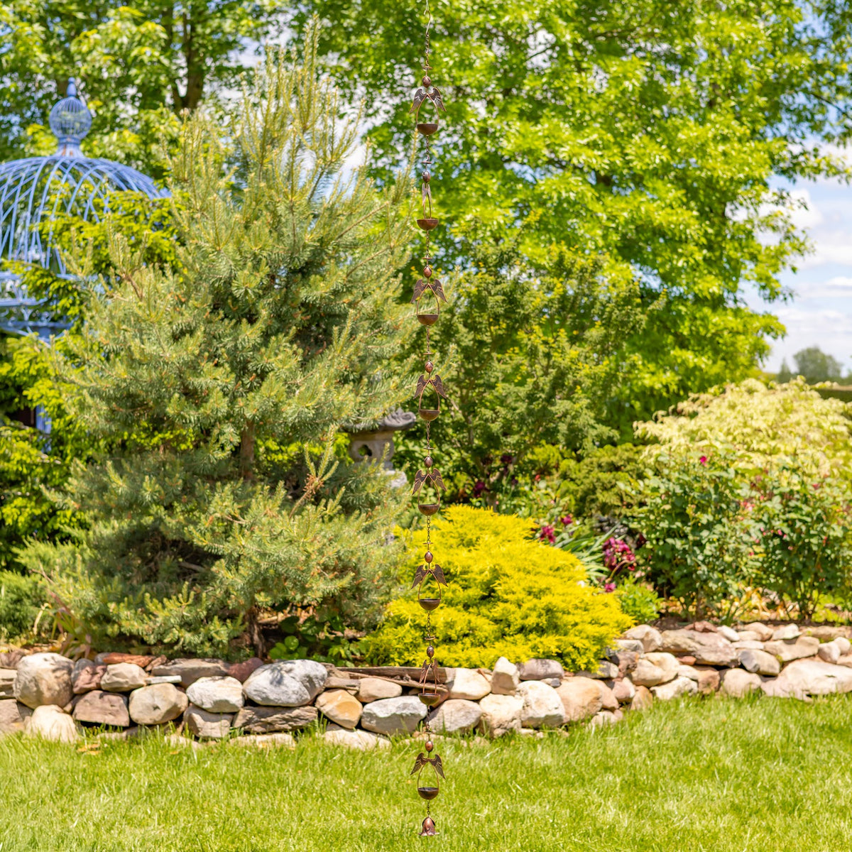 Rain chain hangs from above into a landscaped garden, with trees, shrubs, flowers, and a stone border.