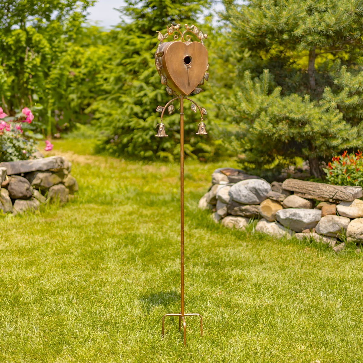 Tall garden stake birdhouse standing in grass, with a heart-shaped body and two hanging bells.