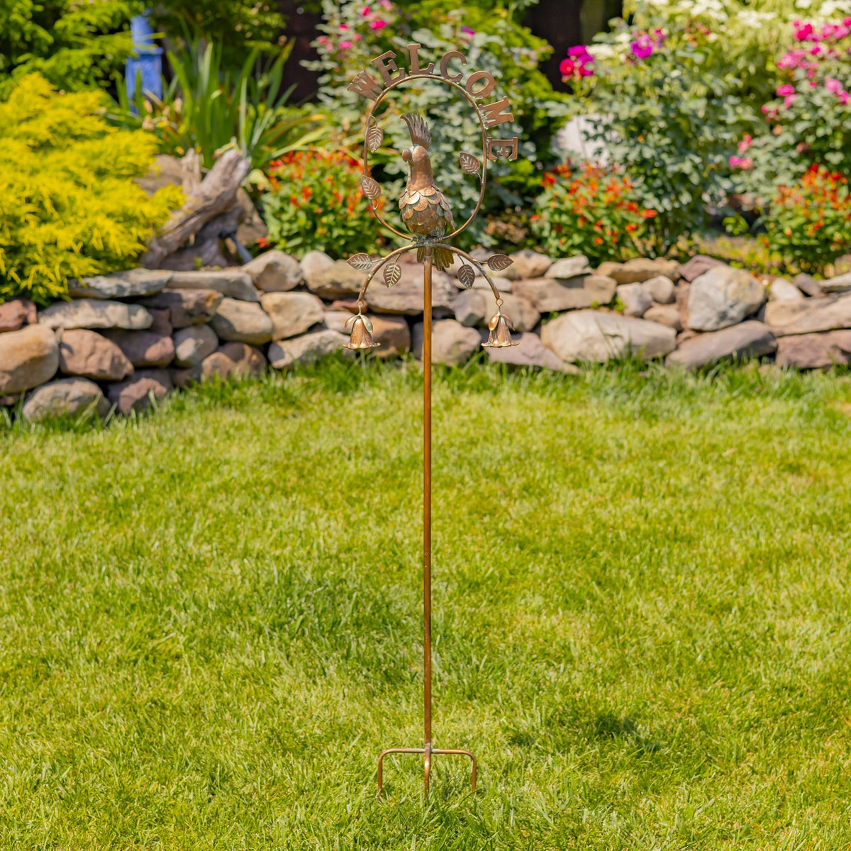 Full garden stake standing in a lawn, with a bird centerpiece, curved frame and bells above the pointed support.