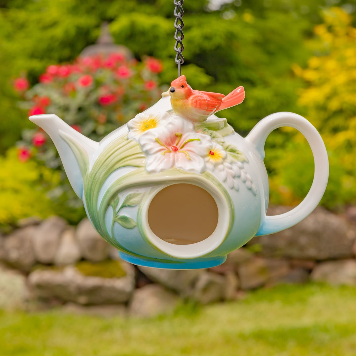 Pale green teapot birdhouse with a red bird and white flower hanging in a garden