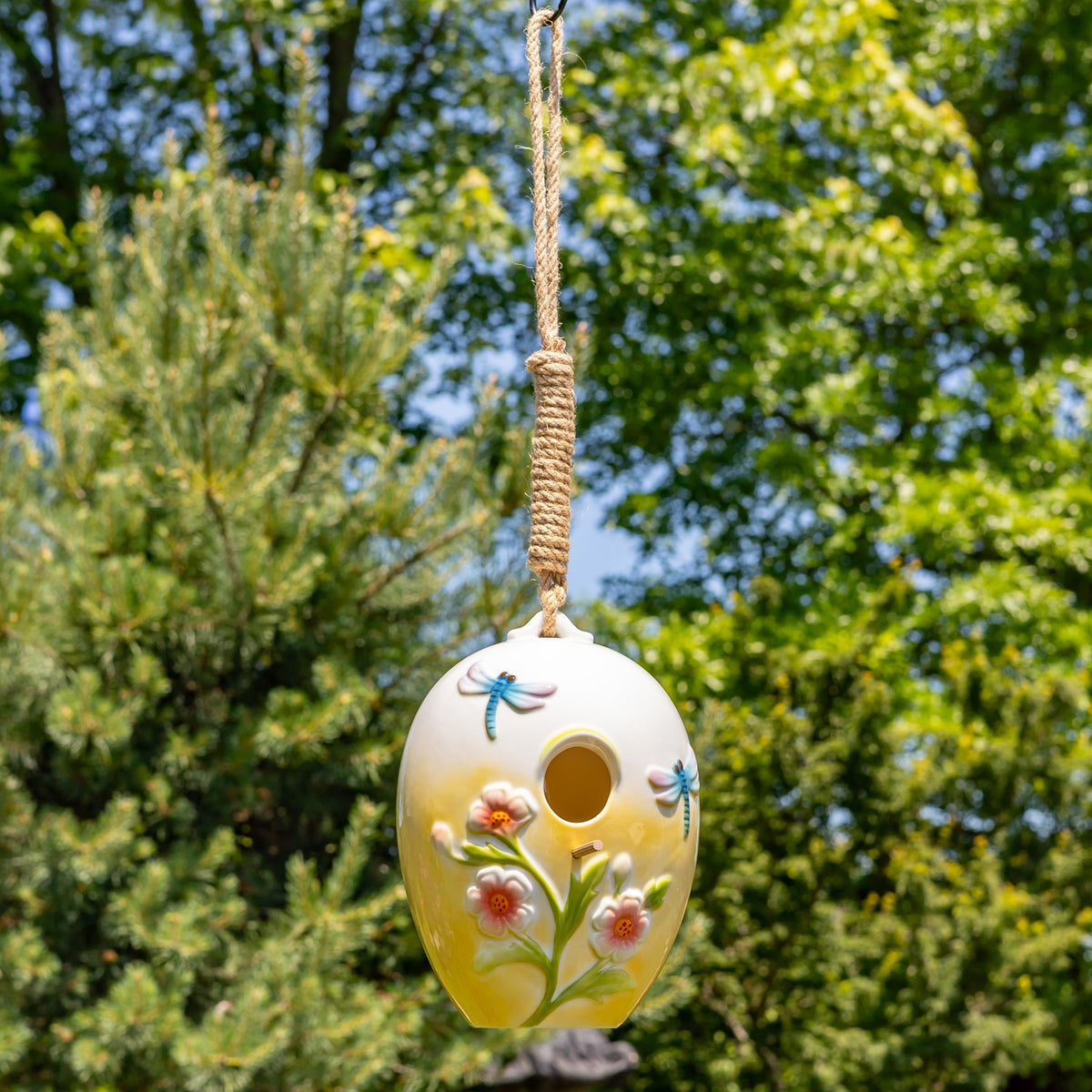 Hanging birdhouse shown at a distance among leafy trees, with its long rope visible above.