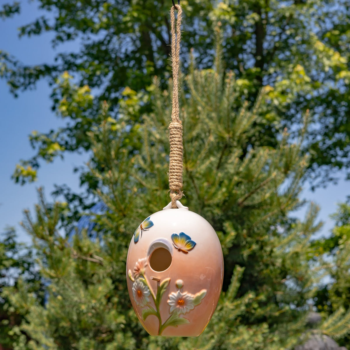 Small floral birdhouse suspended by a long rope in front of a garden tree.