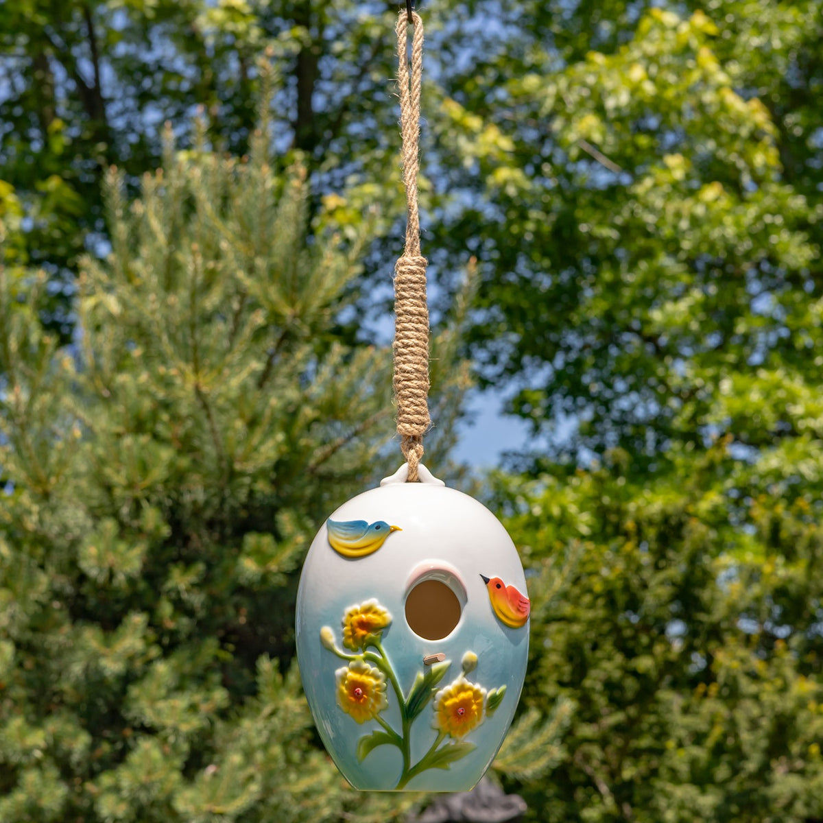 A hanging birdhouse shown at a distance, with its long rope visible against leafy trees.