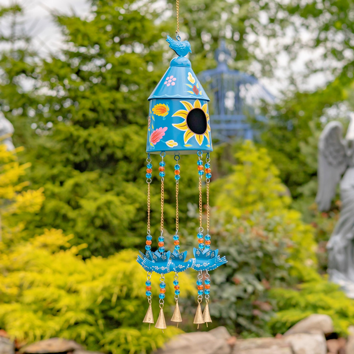 Bright blue birdhouse wind chime with a small bird topper, colorful beads and dangling bells among garden greenery.