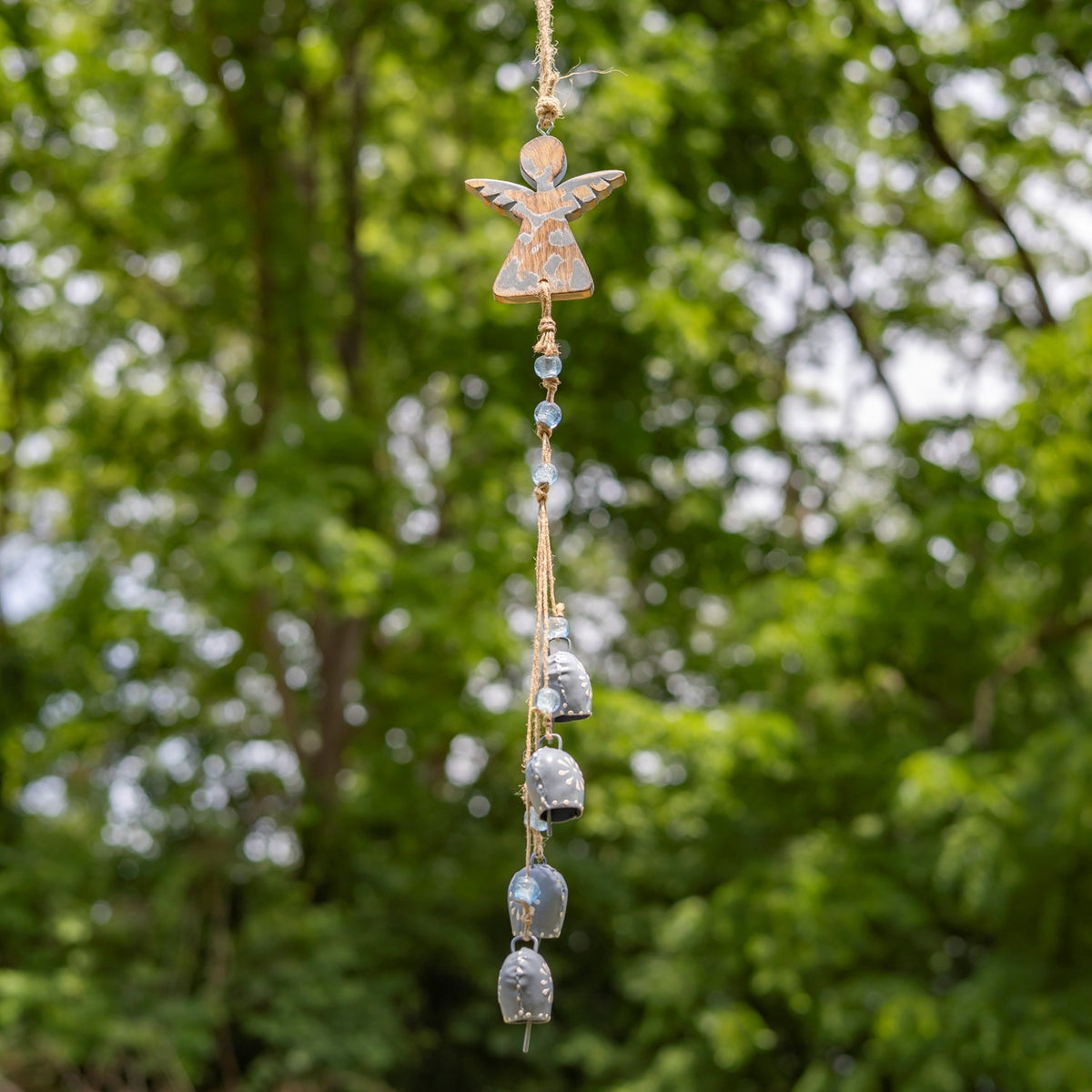 A pale blue angel wind chime with blue beads and silver-gray bells hangs among green trees.