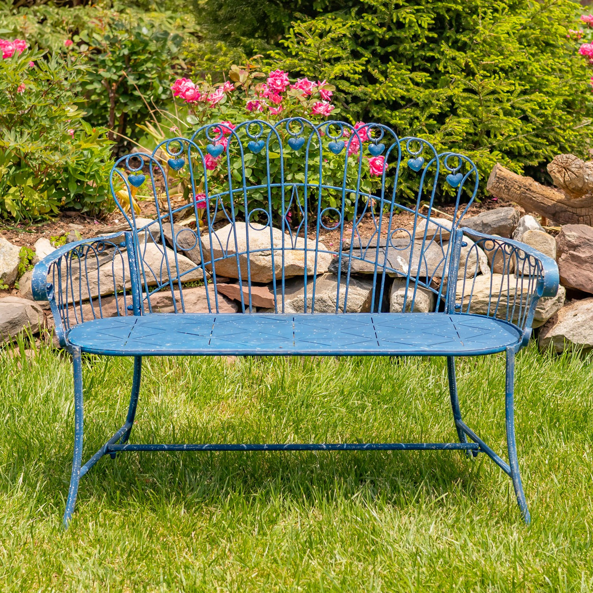 Blue iron garden bench shown straight on in a garden with flowers behind it