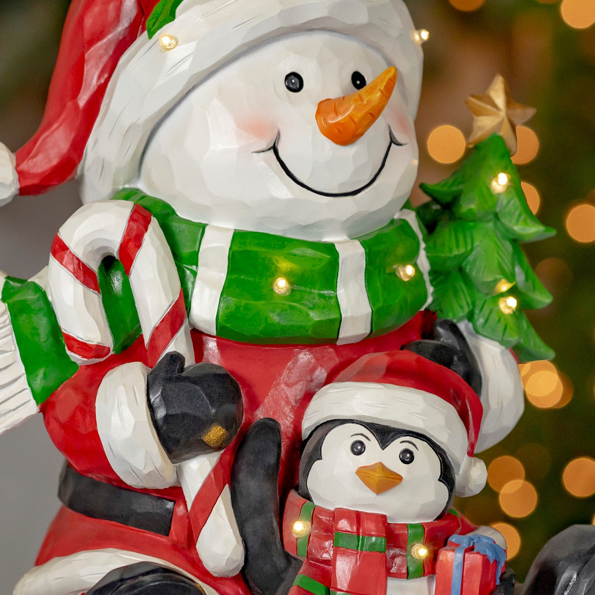 Close view of the snowman's face and scarf, with a penguin holding a wrapped gift beside a glowing tree.