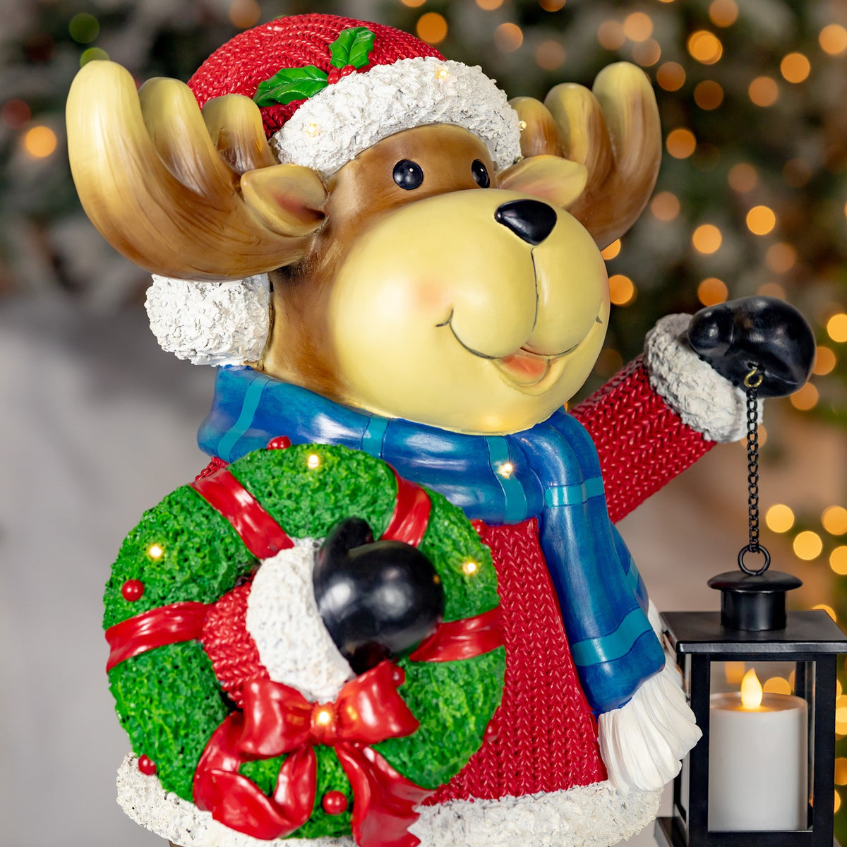 Close view of the moose’s smiling face, blue scarf, wreath, and hanging lantern.